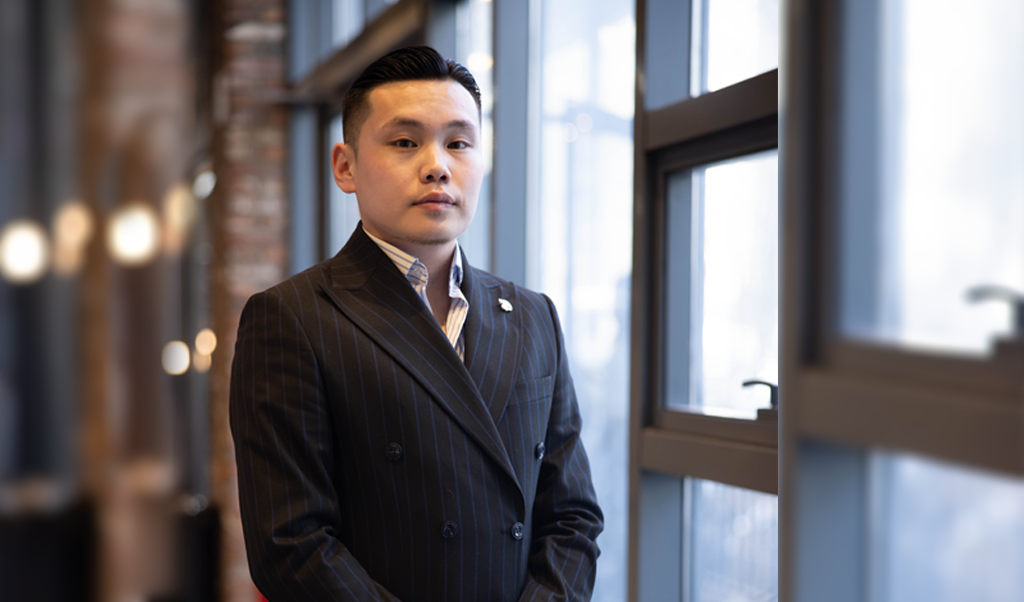 Бараан зураастай костюм өмссөн бизнес эрхлэгч том цонхтой орчин үеийн оффисын орчинд итгэлтэйгээр зогсож байна. Зөөлөн гэрэлтүүлэг, бүдэг арын дэвсгэр нь гялалзсан, мэргэжлийн уур амьсгалыг тодотгож, цэцэглэн хөгжиж буй компанийн орчныг илтгэнэ. - Business.MN