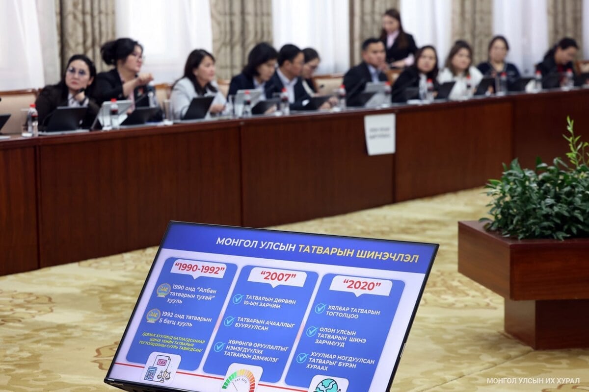 A group of people sits at a long conference table, attentively listening. In the foreground, a screen displays information in Mongolian about tax reforms from 1990 to 2020, using icons and colorful graphics.