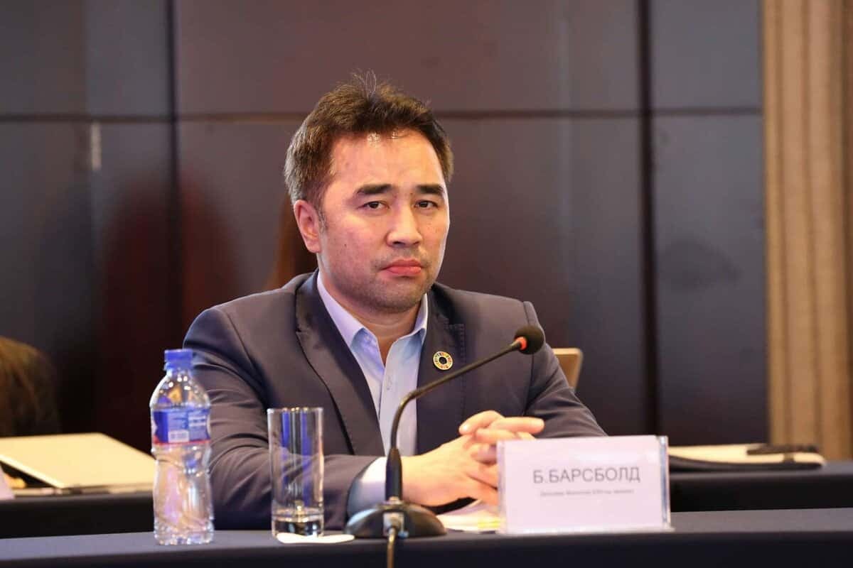 Урд талд нь "Б.Барсболд" гэсэн бичиг наасан хурлын ширээний ард костюмтай эр сууж байна. Тэр ширээн дээр микрофон, аяга ус, усны савтай. Цаана нь бусад оролцогчид, бараан өнгийн том хана байгаа нь энэхүү нөлөө бүхий бизнес эрхэлдэг аж. - Business.MN