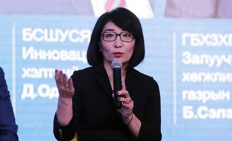 Нүдний шил, хар хувцастай эмэгтэй гараараа дохиж микрофон руу ярьж байна. Тэр кирилл үсгээр бичсэн арын дэвсгэрийн өмнө сууж байна. - Business.MN