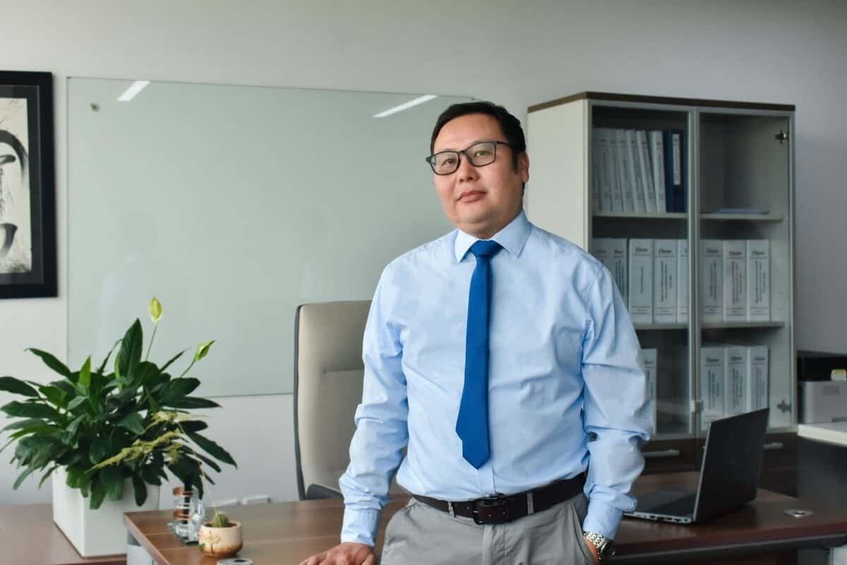 Цэнхэр цамц, зангиа зүүсэн нүдний шил зүүсэн өөртөө итгэлтэй энтрепренер орчин үеийн оффис дээр зогсож байна. Түүний ард цагаан самбар, ургамал, тавиурууд нь цагаан өнгийн холбогчоор дүүрсэн байдаг. Офис нь мэргэжлийн, зохион байгуулалттай дүр төрхтэй. - Business.MN