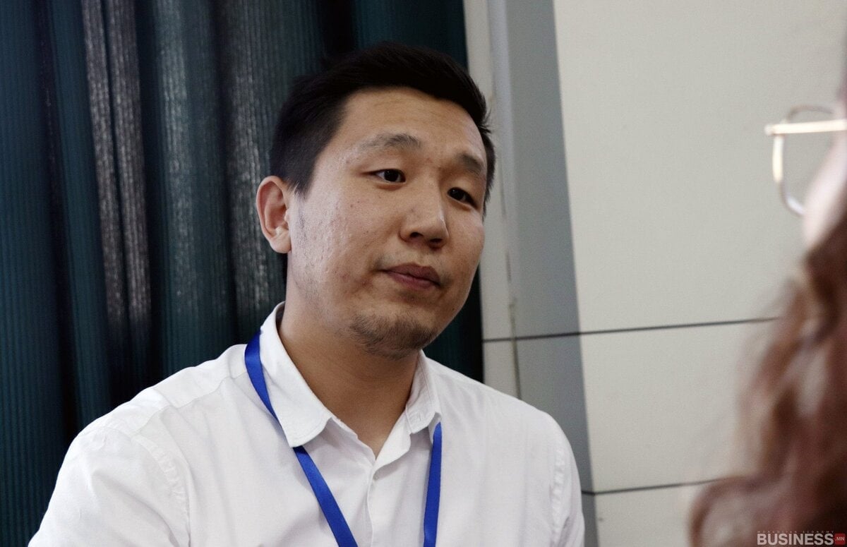 Цагаан цамц, цэнхэр оосор өмссөн эр ярьж байна, баруун талд нь өөр хүний нэг хэсэг харагдаж байна. Арын дэвсгэр нь харанхуй хөшиг, цайвар өнгийн ханыг агуулдаг. - Business.MN