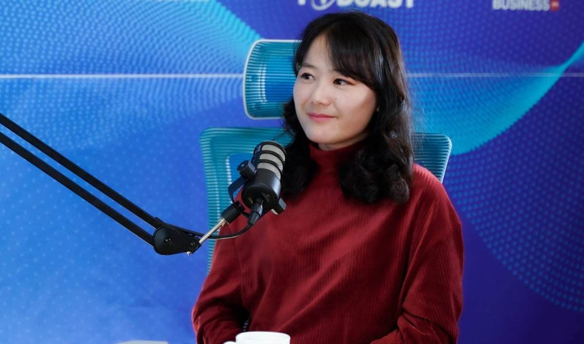 Урт хар үстэй хүн цайвар сандал дээр суугаад индэрт бэхлэгдсэн микрофоноор ярьж байна. Тэд улаан хүзүүвчтэй цамц өмссөн байна. Арын дэвсгэр дээр podcast лого, хийсвэр хээ бүхий цэнхэр өнгийн загвар нь бизнес болон бизнес эрхлэгчдийн талаарх хэлэлцүүлгийг илтгэж байна. - Business.MN