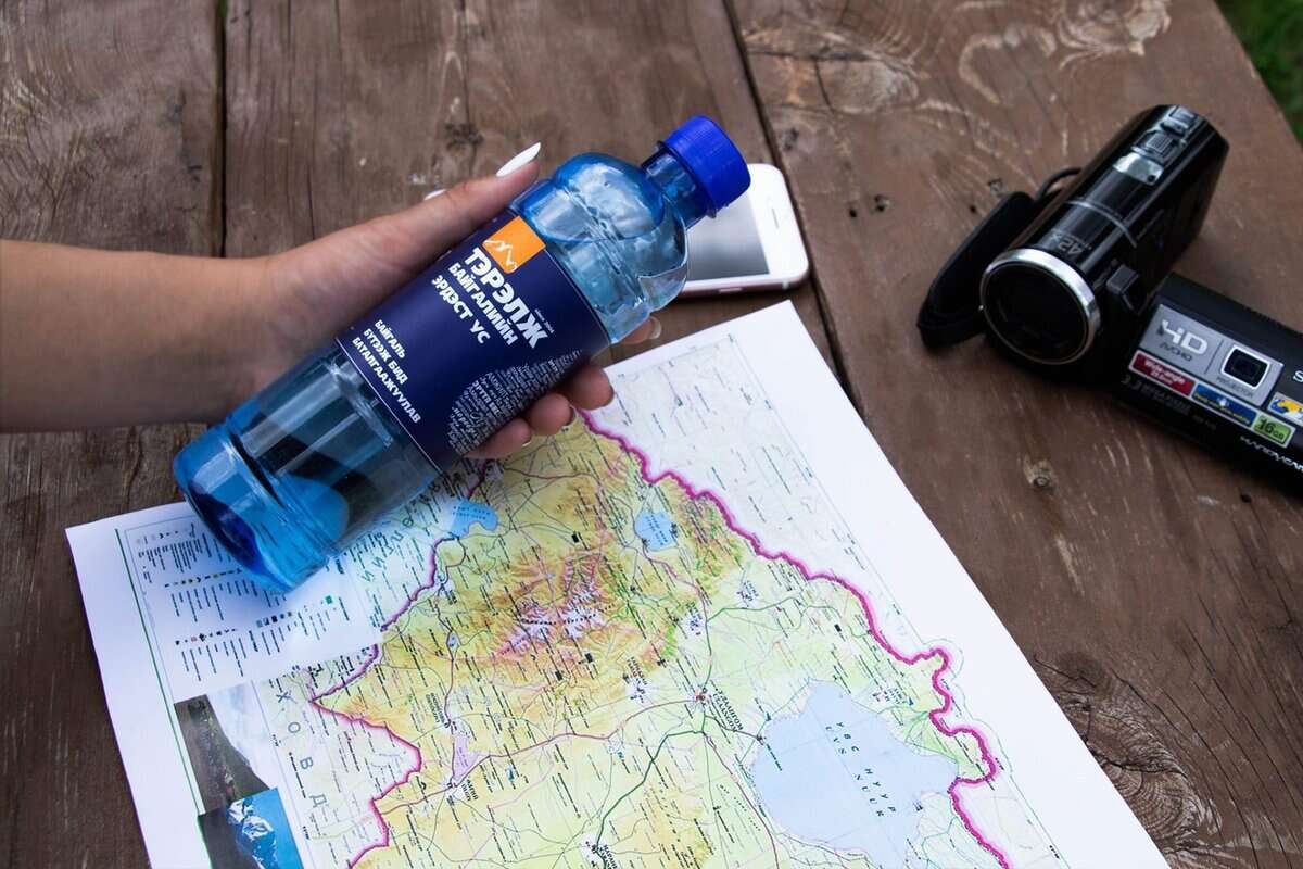 Модон гадаргуу дээр дэлгэсэн нарийвчилсан газрын зураг дээр цэнхэр усны сав барьсан гар. Газрын зургийн хажууд хар камер, цагаан ухаалаг утас байрлуулсан нь энтрепренерийг гадаа адал явдал, аялалд бэлтгэхийг санал болгож байна. - Business.MN