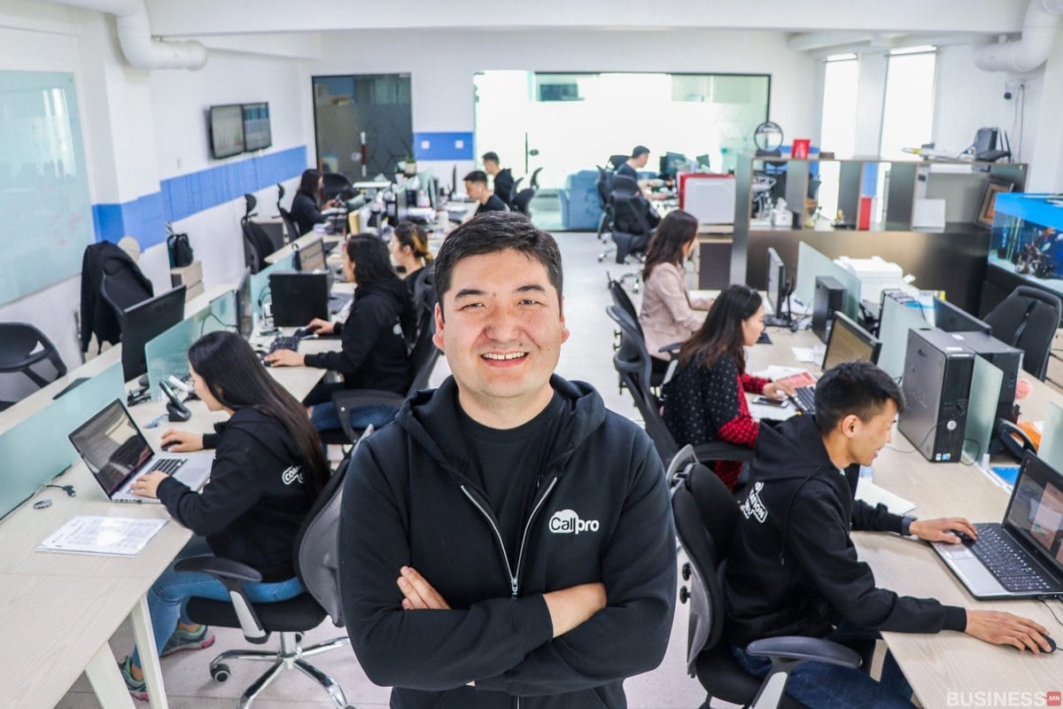 Инээмсэглэсэн энтрепренер албан тасалгаанд гараа зөрүүлэн зогсож байхад цаана нь хэдэн хүн компьютер дээр ажиллаж байна. - Business.MN