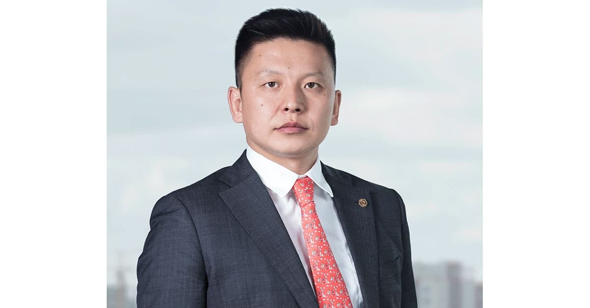 Харанхуй костюм, улаан хээтэй зангиа өмссөн эрэгтэй цайвар дэвсгэр дээр зогсож байна. Богино хар үстэй, төвийг сахисан царайтай. Зураг нь мэргэжлийн хөрөг зургийн хэв маягийг илэрхийлдэг. - Business.MN