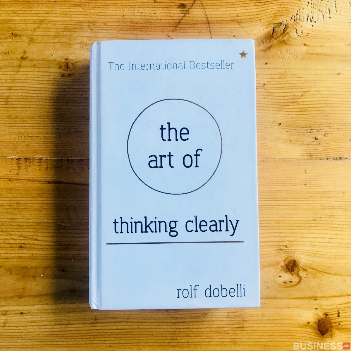 Рольф Добеллигийн "Тодорхой сэтгэх урлаг" хэмээх номыг модон гадаргуу дээр байрлуулсан. Хавтас нь энгийн, цайвар дэвсгэр дээр хар тексттэй, баруун дээд буланд жижиг одтой. - Business.MN