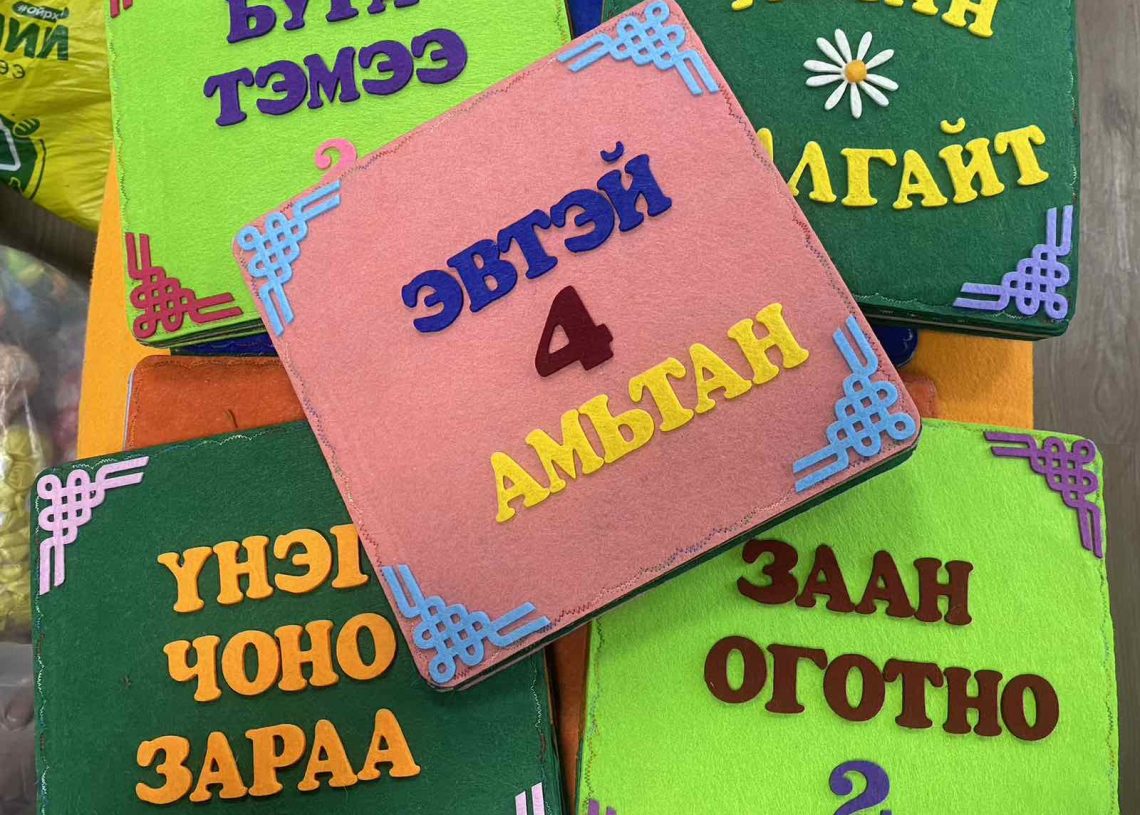 Colorful felt squares with Mongolian text and numbers are displayed. Each square has a unique border design. The center square reads "ЭВТЭЙ 4 АМЬТАН" and is surrounded by squares labeled with numbers and animal names.