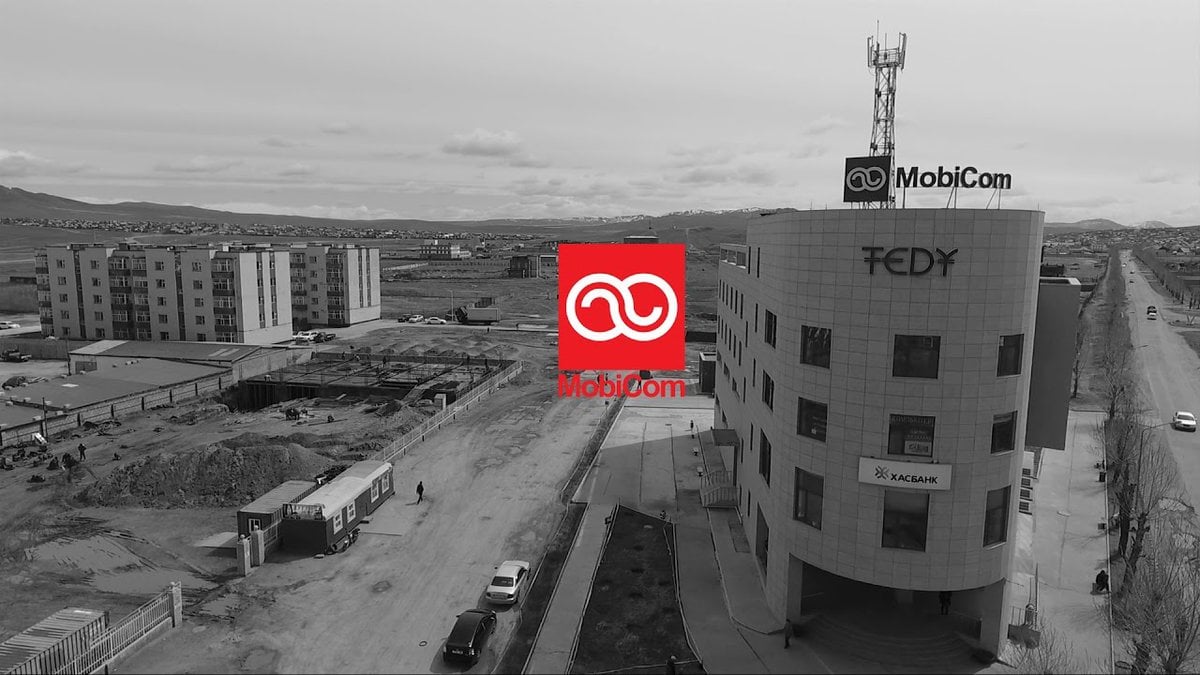 "Теди", "Мобиком" лого бүхий орчин үеийн барилгын саарал өнгийн агаараас. Энэхүү тохиргоонд орон сууцны барилга, барилгын талбай, үүлэрхэг тэнгэрийн дор алс хол руу чиглэсэн зам багтана. - Business.MN