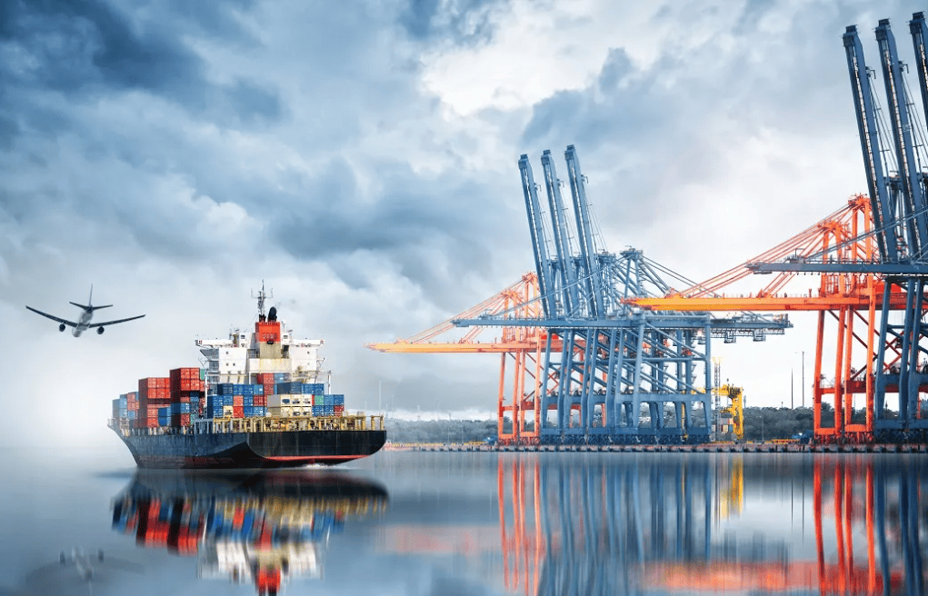 A cargo ship laden with colorful containers sails near a large port with towering cranes. An airplane flies in the cloudy sky above, reflecting on the calm water, creating a dynamic industrial scene.