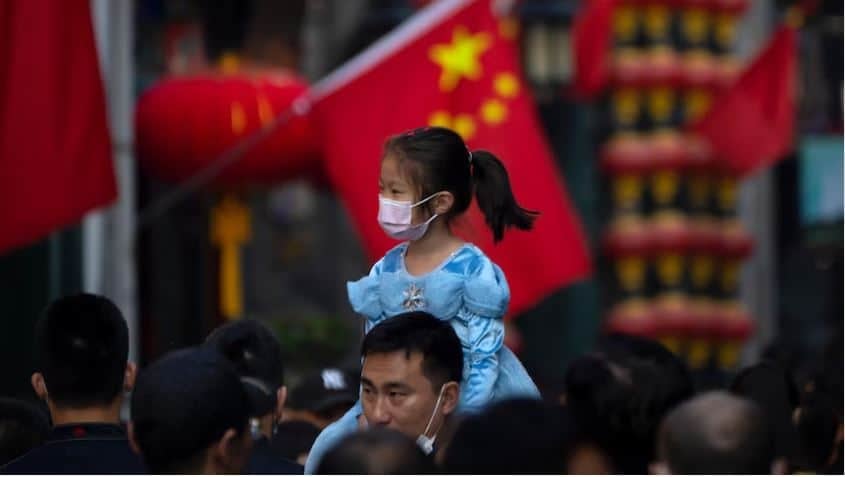 Цэнхэр даашинз, маск зүүсэн залуу охин олон хүнтэй гадаа эрэгтэй хүний мөрөн дээр сууж байна. Ар талд нь улаан дэнлүү, Хятадын тугнууд харагдаж байгаа нь баяр ёслол, соёлын арга хэмжээг илтгэнэ. - Business.MN