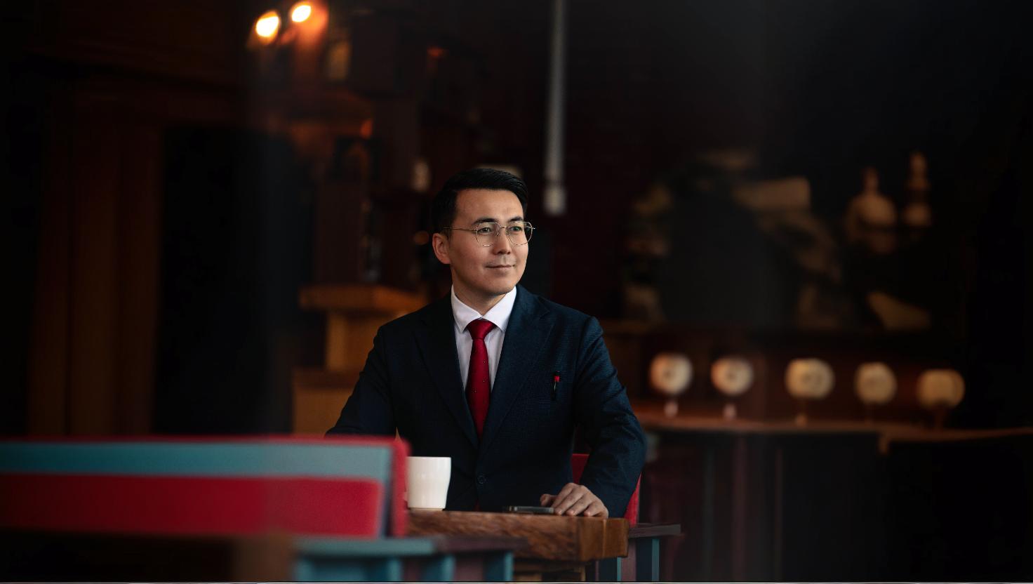 A man in a suit and red tie sits at a wooden table in a dimly lit room. He is holding a white cup and facing forward, with a thoughtful expression. The background is softly blurred, highlighting warm lighting and subtle decor.