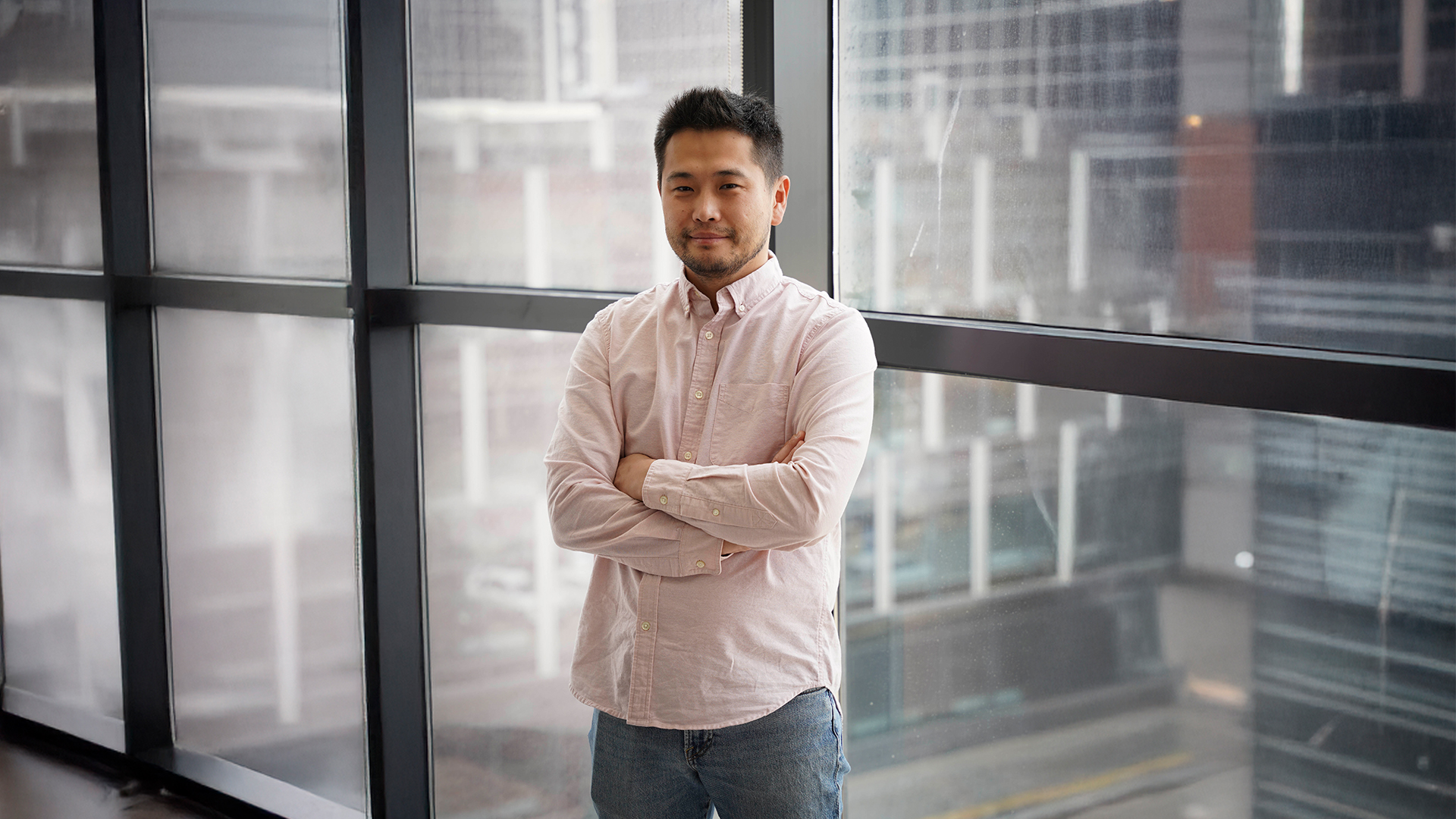 A man in a light pink button-up shirt and jeans stands with arms crossed, looking at the camera. Indoors, large windows reveal an urban cityscape—технологийн дэвшил чанартай үйлчилгээг хослуулна илтгэнэ.