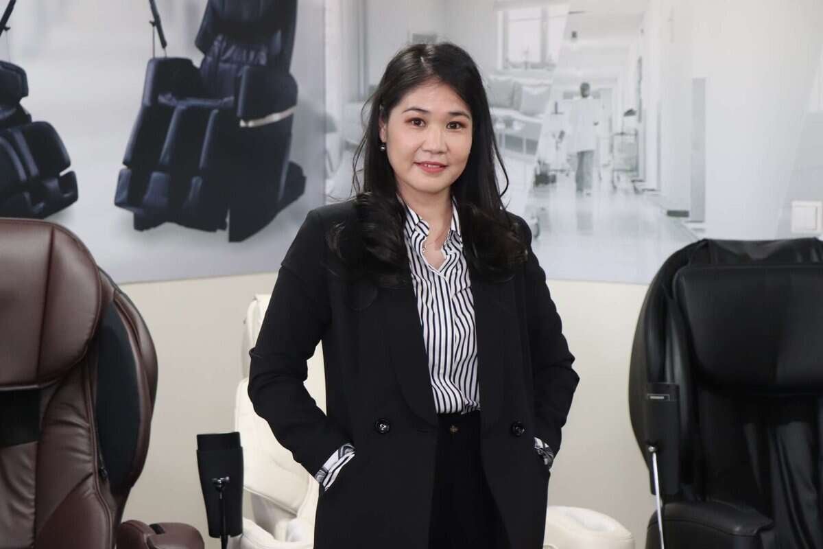 Хар пиджак өмссөн эмэгтэй хэд хэдэн массажны сандал дунд итгэлтэй зогсож байна. Тэр урт хар үстэй, судалтай цамц өмссөн байна. Арын дэвсгэр нь орчин үеийн ажлын байрны дүр төрхийг харуулж байна. - Business.MN