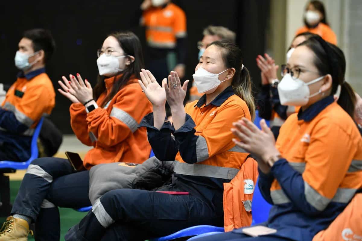 Улбар шар өнгийн хүрэм, маск өмссөн хэсэг хүмүүс цэнхэр сандал дээр суугаад алга ташин сууна. Тэд бизнес эрхлэгчийн эзэмшдэг агуулах эсвэл үйлдвэрлэлийн байгууламж байж магадгүй ажлын байранд байгаа бололтой. Зарим нь хамгаалалтын хантааз, нүдний шил зүүсэн байна. Тэдний илэрхийлэл нь тэднийг оролцож байгаа эсвэл талархаж буйг илтгэнэ. - Business.MN