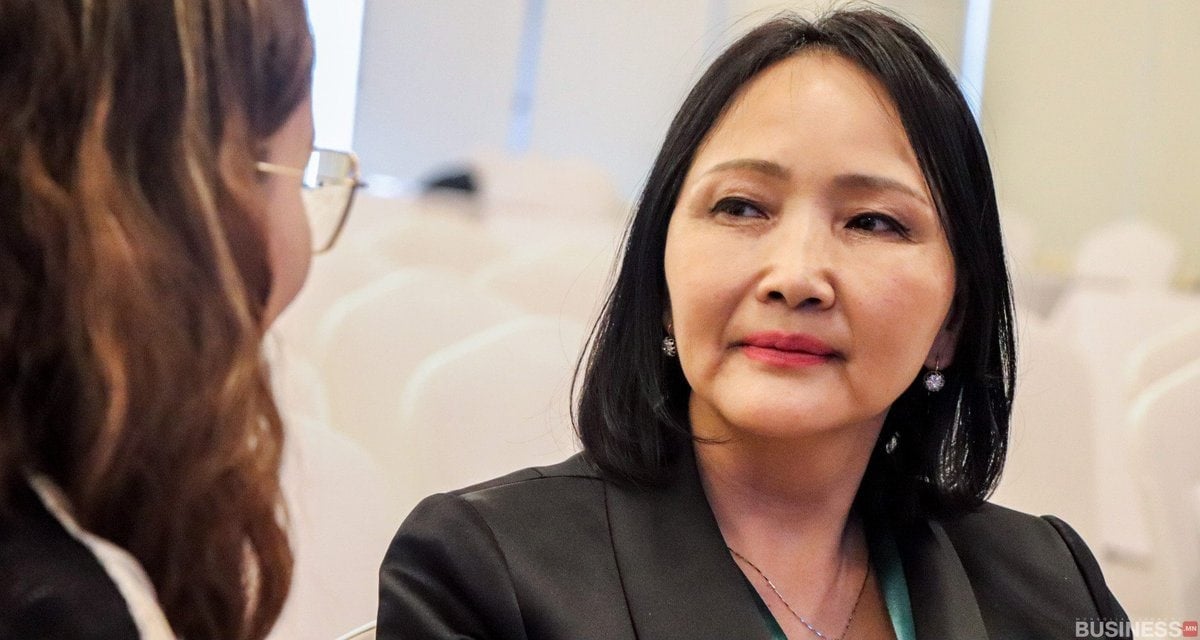 Хоёр эмэгтэй сууж, ярилцаж байна. Нэг нь урт хар үстэй, хар пиджак өмсдөг бол нөгөө нь буржгар үстэй, нүдний шилтэй нь хажуу талаас нь харагдана. Тэд арын дэвсгэр дээр бүдгэрсэн сандал бүхий мэргэжлийн орчинд байгаа бололтой. - Business.MN