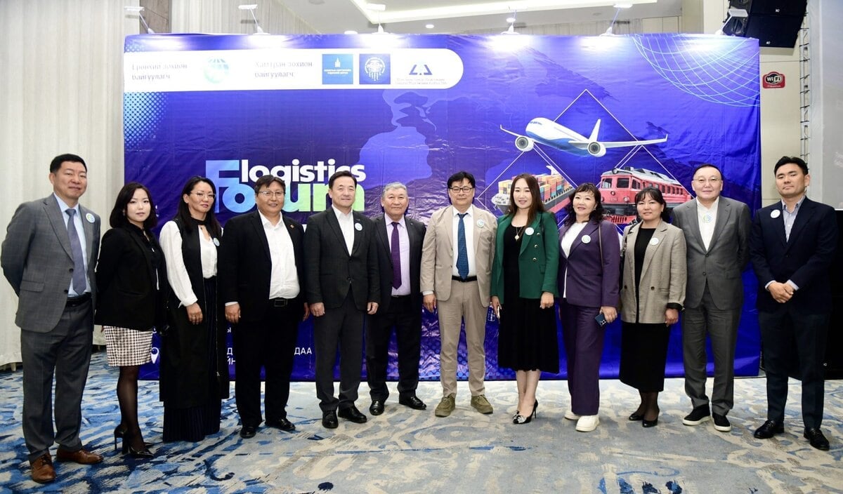 A group of thirteen people, dressed in business attire, stand in a row smiling in front of a blue "Logistics Forum" backdrop featuring images of an airplane, helicopter, and cargo ship.