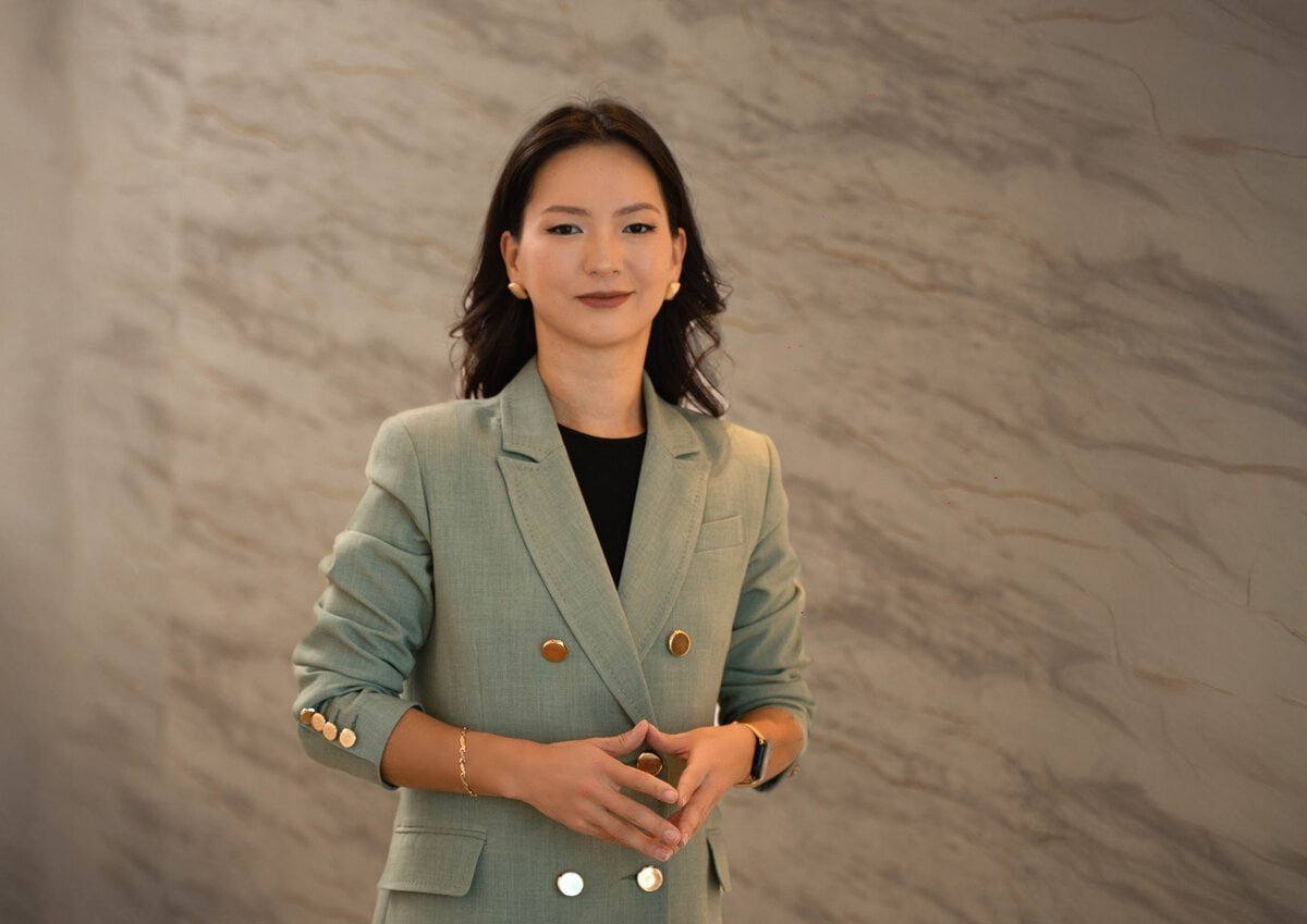 A person with shoulder-length hair stands confidently against a marble-patterned wall. They are wearing a light green double-breasted blazer with rolled-up sleeves. Their hands are placed together in front of them.