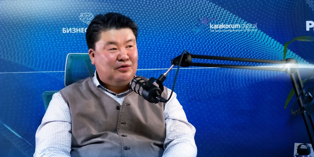 Нэг хүн микрофоны өмнө суугаад подкаст эсвэл ярилцлага хийж байгаа бололтой. Тэрээр цайвар өнгийн цамц, саарал хантааз өмссөн энтрепренер юм. Арын дэвсгэр дээр кирилл үсгээр бичсэн зарим бичвэртэй цэнхэр, ногоон дижитал хээ, "karakorum digital" логотой. - Business.MN