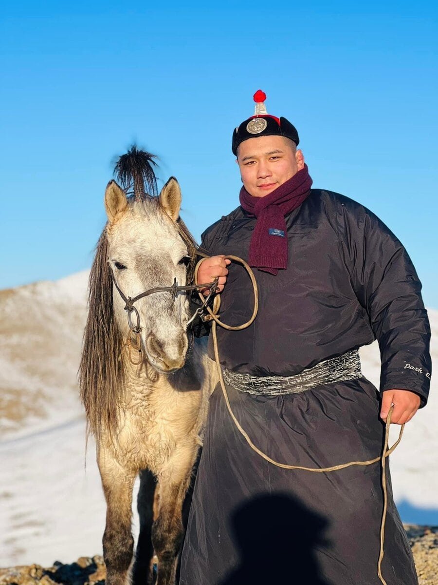 A man in traditional Mongolian clothing stands outdoors holding the reins of a gray horse, with snow-covered mountains and a clear blue sky in the background.