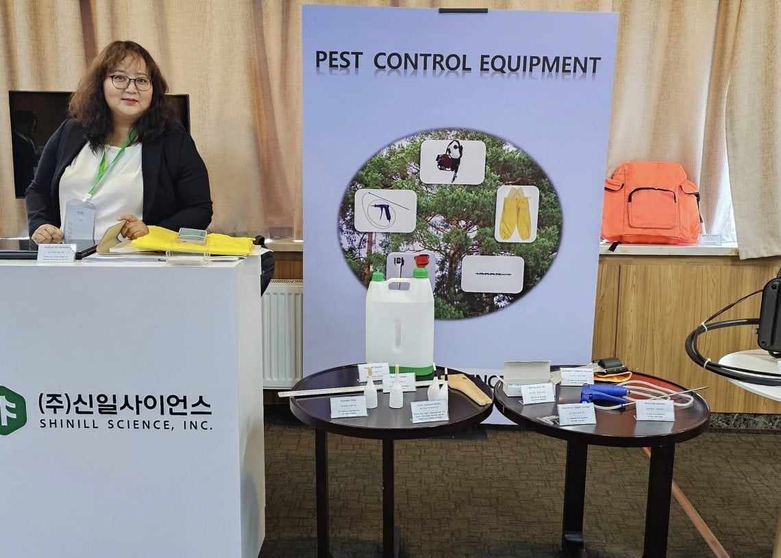 A woman stands at a booth displaying pest control equipment. The booth features a sign with pest illustrations, various pest control tools on tables, and a banner labeled "SHINIL SCIENCE, INC.
