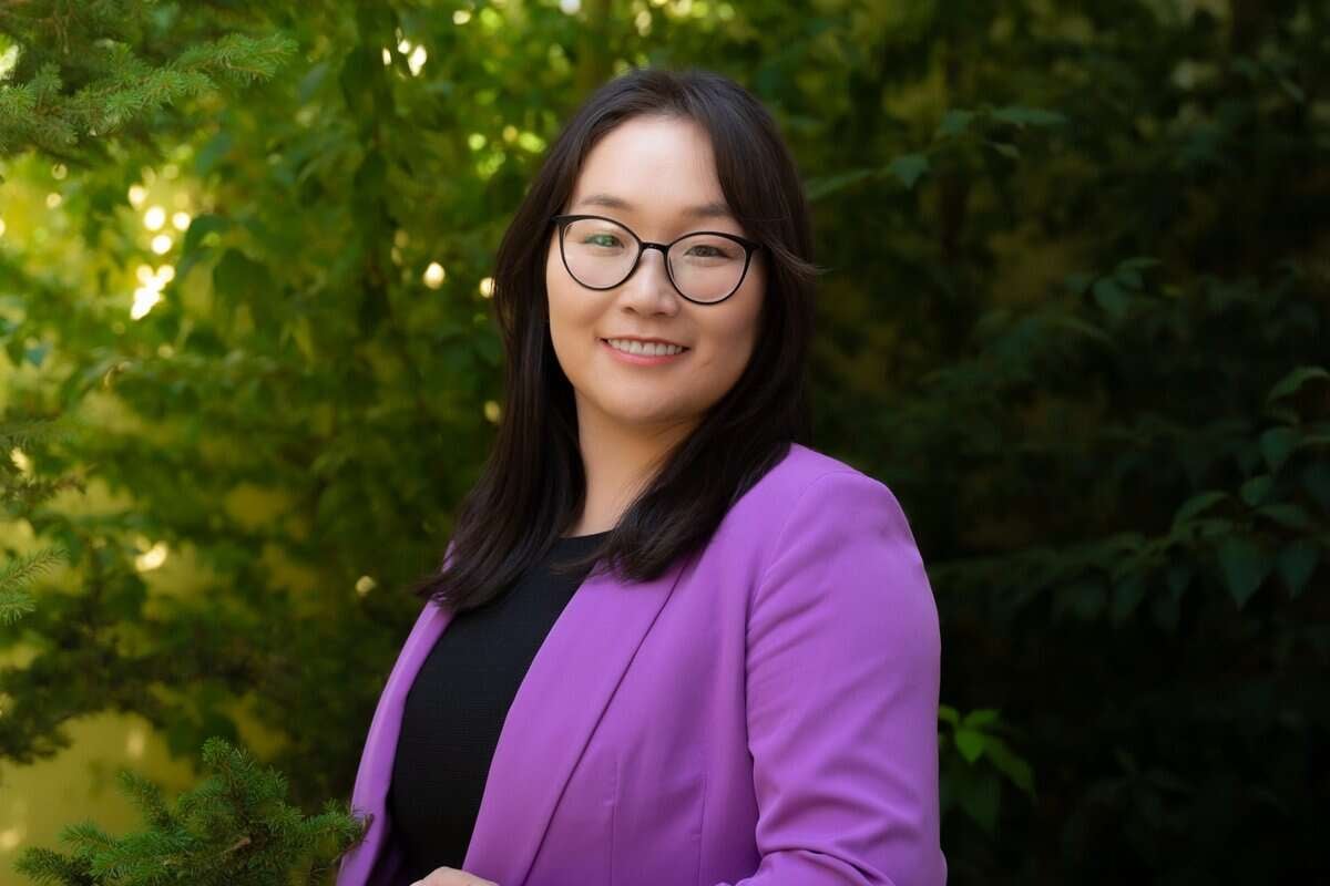 Урт үстэй, нүдний шилтэй хүн инээмсэглэж, хувцасны мэдрэмжийг харуулж байна. Тэд хар орой дээр нил ягаан өнгийн пиджак өмсөж, гадаа өтгөн ногоон навчис дунд зогсож байна. - Business.MN