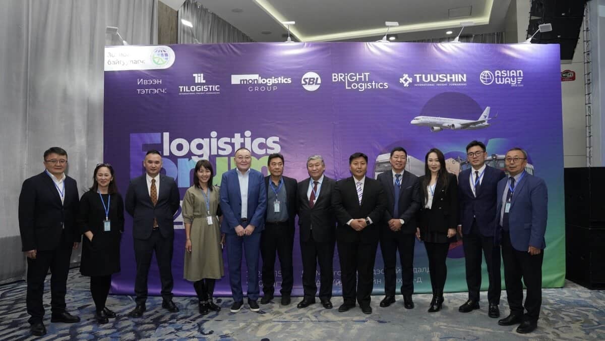 A group of people stands in front of a purple banner at the Монголын Тээвэр Зуучлагчдын Нэгдсэн Холбоо ‘Ложистикс Форум 2024’. They are dressed in formal attire, smiling at the camera, with a model airplane featured on the banner.