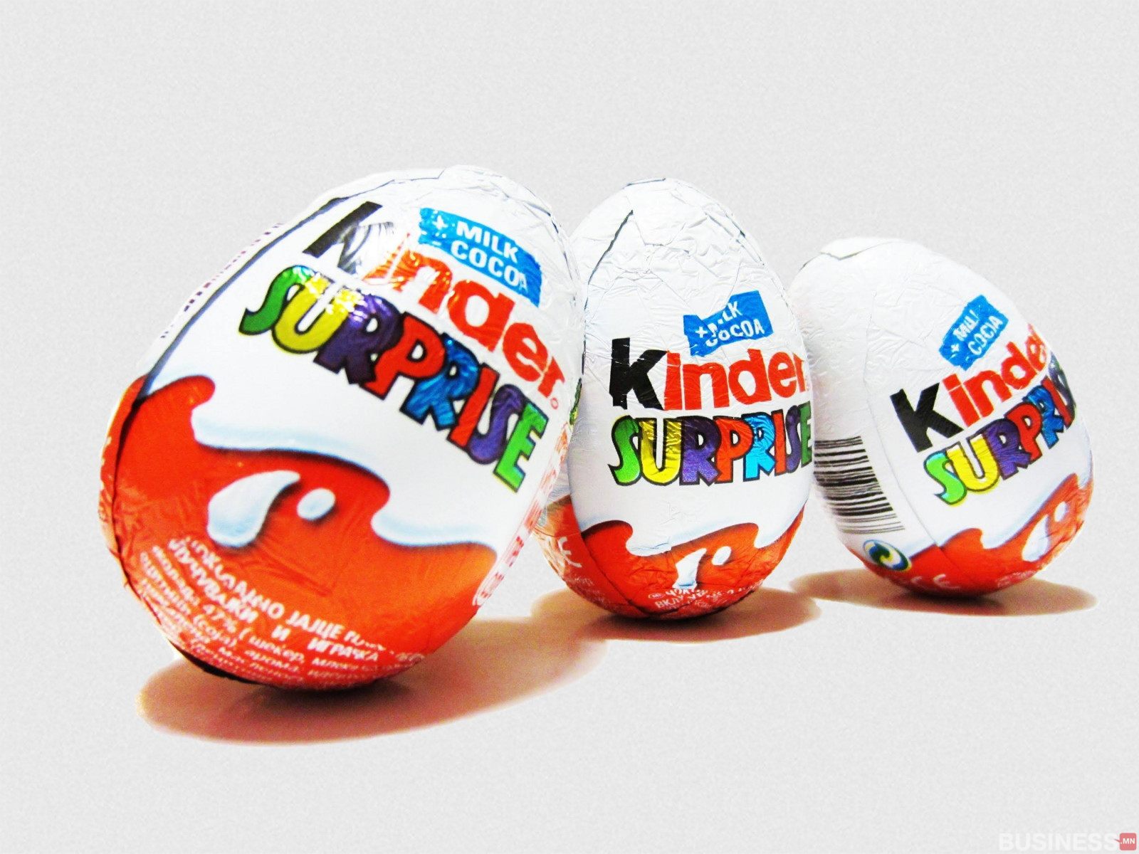 Өнгө өнгийн боодолтой гурван Kinder Surprise өндөг нь энгийн дэвсгэр дээр босоо зогсож байна. Боодол дээр брэндийн лого, гялалзсан улаан өнгөтэй, "Milk Cocoa. - Business.MN