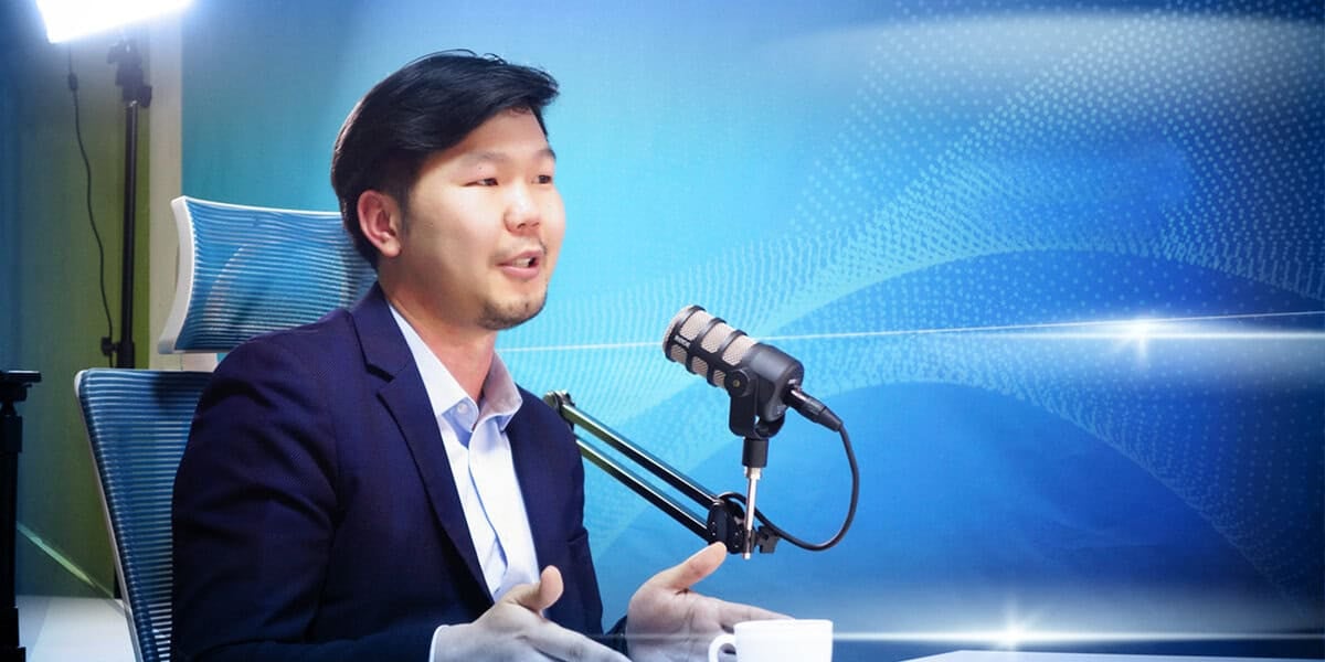 Ажил хэрэгч хувцастай эр микрофоны өмнө суугаад ярьж байна. Арын дэвсгэр нь эрч хүчтэй, хийсвэр цэнхэр дизайнтай, мэргэжлийн гэрэлтүүлэгтэй. Бизнесийн энтрепренер гараараа дохиж, хөдөлгөөнтэй, илэрхий харагдаж байна. - Business.MN