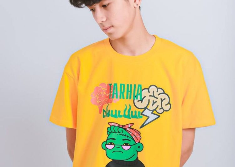 Ногоон царай, аянга цахилгаантай хүүхэлдэйн киноны загвар бүхий тод шар өнгийн график подволк өмссөн буржгар хар үстэй залуу энгийн цайвар дэвсгэр дээр зогсож, доошоо харж байна. - Business.MN