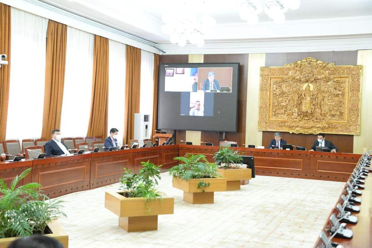 Таван бизнес эрхлэгч хагас дугуй хэлбэртэй ширээтэй том хурлын танхимд сууж байна. Виртуал уулзалтад оролцогчдыг харуулсан гурван том дэлгэцийг хананд суурилуулсан байна. Өрөөнд цагаан хөшиг, бор өнгийн хөшиг бүхий том цонхтой. Саванд ургамлыг төвд байрлуулна. - Business.MN