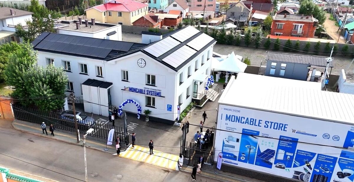 Дээвэр дээрээ нарны зай хураагууртай, орцны дээгүүр цаг зүүсэн, чимэглэсэн нуман хаалгатай цагаан ногоон байгууламжийн агаараас. Хажууд нь "МОНКАБЛ ДЭЛГҮҮР"-ийн том майхан, эргэн тойронд цугласан хүмүүс, хажууд нь зэргэлдээх байшингууд харагдана. - Business.MN