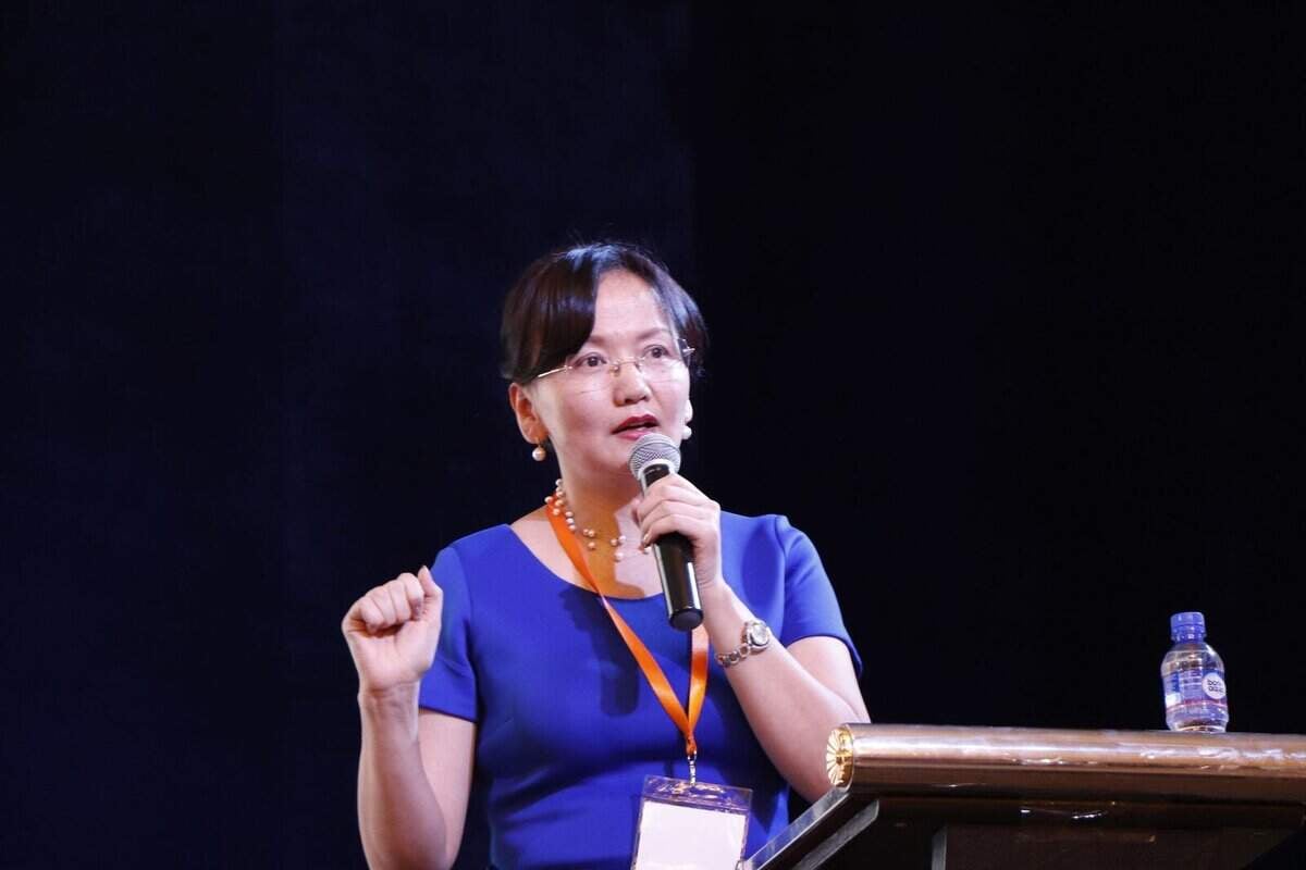 Цэнхэр даашинзтай эмэгтэй индэр дээр зогсож байхдаа микрофоноор ярьж байна. Тэр нүдний шил, улбар шар өнгийн оосор зүүж, индэр дээр усны сав байна. Арын дэвсгэр нь бараан өнгөтэй байна. - Business.MN