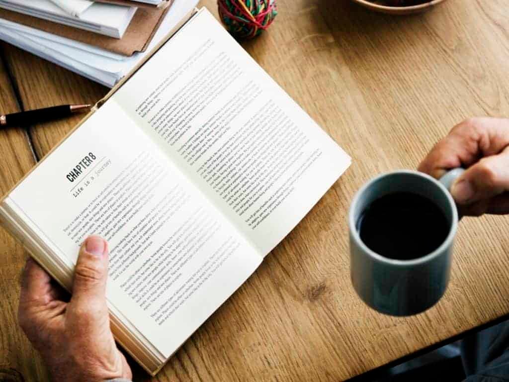 Энтрепренер модон ширээн дээр аяга кофе бариад ном уншиж байна, эргэн тойронд нь тараагдсан цаас, үзэг. - Business.MN