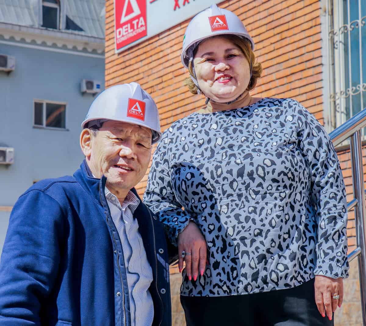 Цагаан малгай өмссөн хоёр хүн тоосгон фасадтай барилгын өмнө зогсож байна. Зүүн талд байгаа хүн хөх өнгийн хүрэм, баруун талд байгаа хүн хээтэй цамц өмсдөг. Тэдний ард байрлах барилга дээр "Дельта" гэсэн тэмдэг харагдана. - Business.MN