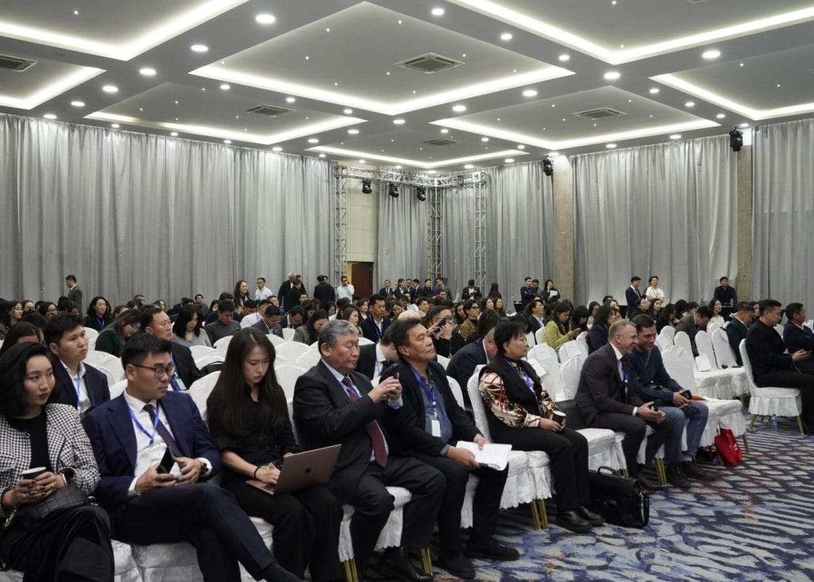 The Монголын Тээвэр Зуучлагчдын Нэгдсэн Холбоо's 'Ложистикс Форум 2024' fills a large conference room with attendees in formal attire, busily using laptops and taking notes. The well-lit space features a patterned carpet and white chairs, providing a professional setting for the event.