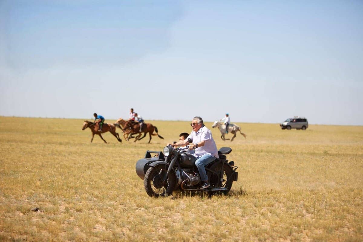 A person rides a motorcycle with a sidecar across a grassy plain. In the background, four people on horseback and an SUV from Номадик Экспедишнс traverse the landscape under a clear blue sky, capturing the adventurous spirit of Жалса Урубшуров.