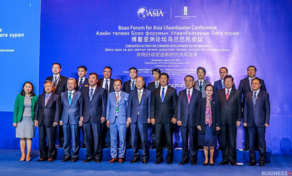 Боаогийн Азийн форумын Улаанбаатар чуулга уулзалтын үеэр албан ёсны хувцастай хэсэг хүмүүс тайзан дээр зургаа авахуулж байна. Тэдний ард олон хэл дээрх бичвэр бүхий цэнхэр дэвсгэр байна. - Business.MN
