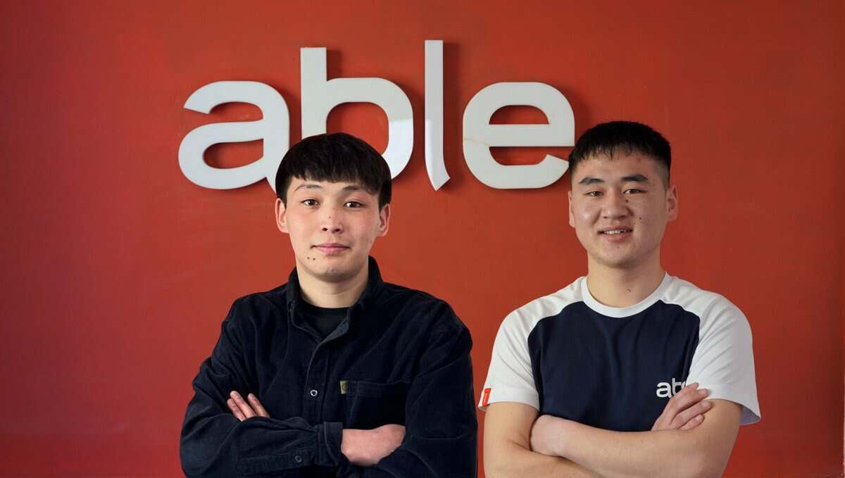 Хоёр энтрепренер залуу улаан хананы өмнө зогсож, ард нь том цагаан үсгээр "чадвартай" гэсэн бичиг бичжээ. Тэд хоёулаа хар үстэй, инээмсэглэж байна. Зүүн талынх нь хар цамц, баруун талынх нь "чадвартай" гэсэн бичигтэй цагаан бараан өнгийн цамц өмсдөг. - Business.MN