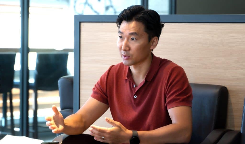 Богино хар үстэй, улаан поло цамц өмссөн эр орчин үеийн оффисын орчинд ширээний ард сууж байна. Тэрээр компанийхаа тухай халуун сэтгэлээр ярихдаа гараараа дохиж, ярианд идэвхтэй оролцдог. Арын дэвсгэр нь цайвар модон хавтан, том цонхтой. - Business.MN