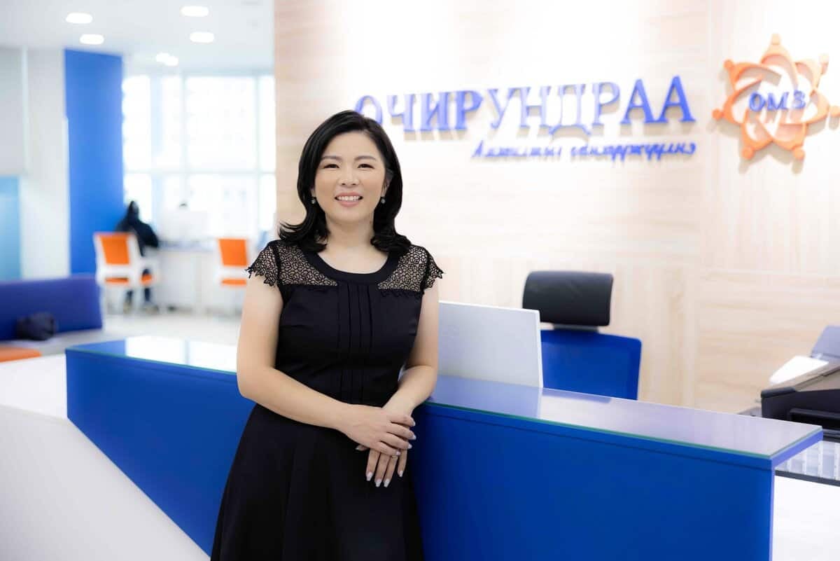 Орчин үеийн оффисын орчинд хар даашинзтай эмэгтэй инээмсэглэн зогсож байна. Түүний ард хүлээн авалтын ширээ, хананд кирилл бичигтэй самбар байдаг. Цэнхэр, цагаан өнгийн гоёл чимэглэлийн элементүүдээр цэцэглэн хөгжиж буй бизнес эрхлэгчдийн мэргэжлийн уур амьсгалыг тусгасан гэрэл гэгээтэй, мэргэжлийн дизайнтай болсон. - Business.MN