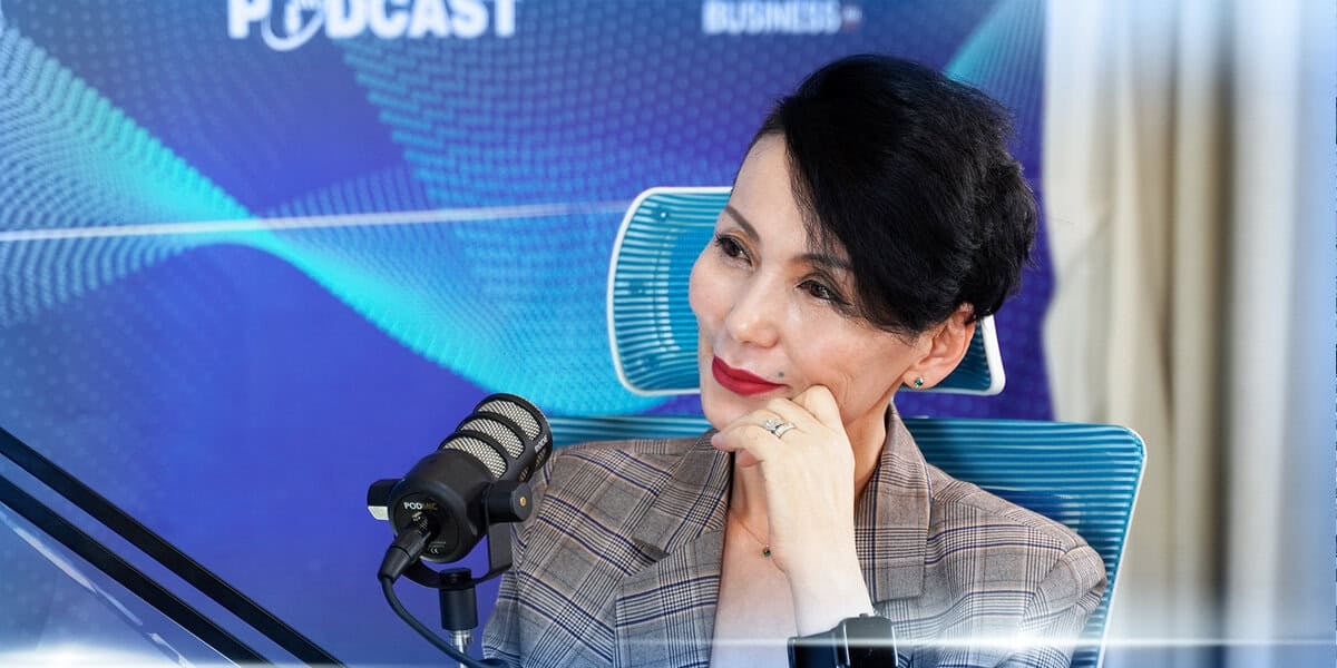 Богино хар үстэй, дөрвөлжин пиджак өмссөн хүн цэнхэр сандал дээр суугаад микрофонд бизнесийнхээ талаар ярьж байна. Тэдний ард подкаст лого, долгионы дизайны элементүүдтэй цэнхэр өнгийн фон байдаг. Энтрепренер гараа эрүүгээ наан бодлогоширон инээмсэглэж байна. - Business.MN