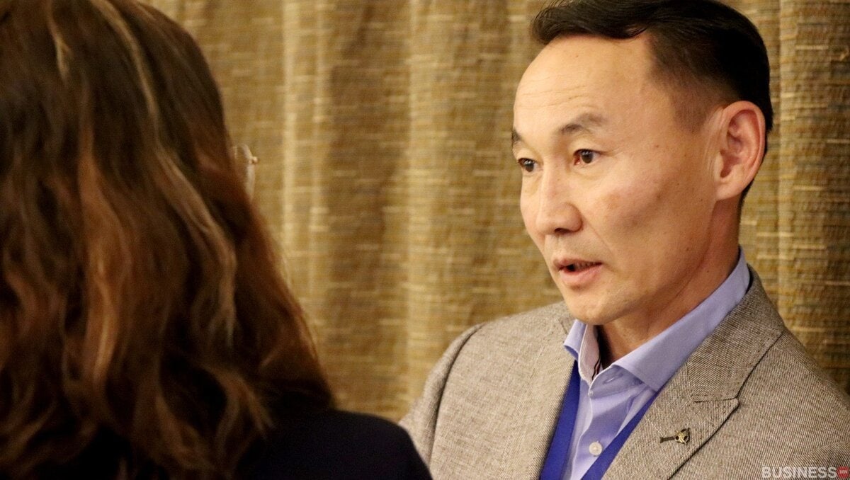 Саарал пиджак өмссөн эрэгтэй нуруу нь камерт байгаа эмэгтэйтэй ярилцаж байна. Арын дэвсгэр нь бүтэцтэй, алтан хүрэн хөшигтэй. - Business.MN