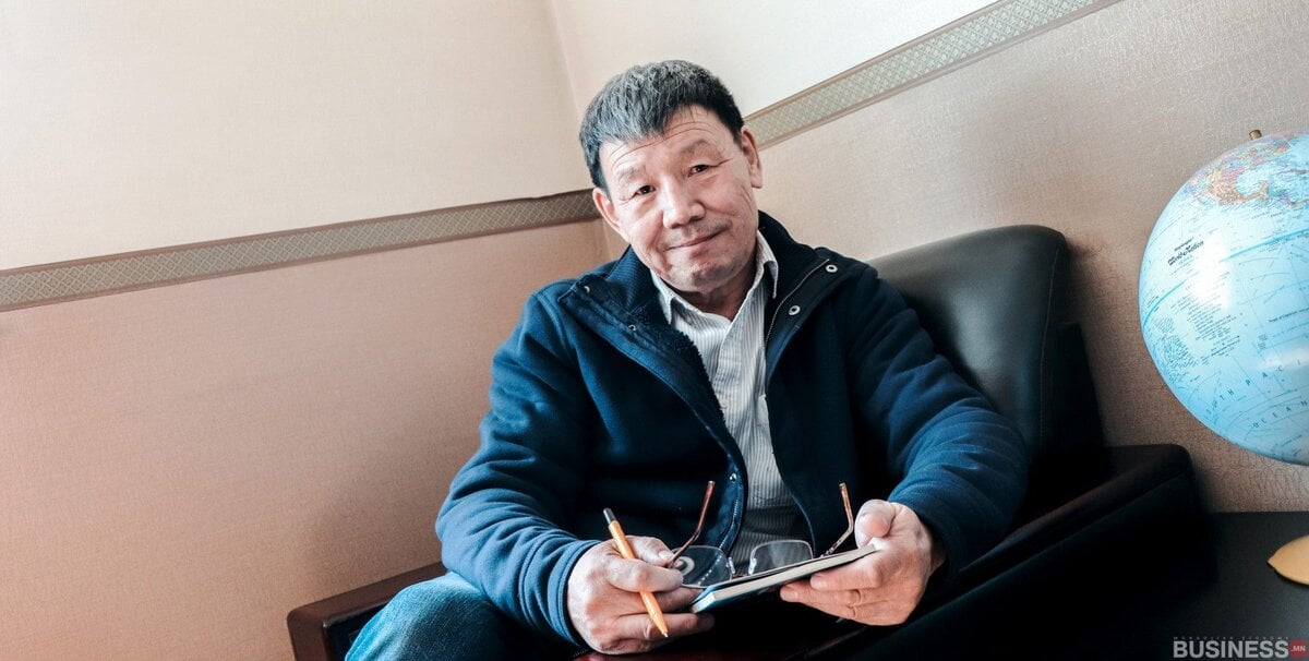 Хөгшин эр түшлэгтэй сандал дээр сууж, үзэг, таблет барьсан. Тэр цэнхэр хүрэм, цагаан цамц өмсдөг. Түүний дэргэд ширээн дээр бөмбөрцөг байдаг. Өрөө нь шаргал ханатай бөгөөд дулаан уур амьсгалыг бүрдүүлдэг. - Business.MN