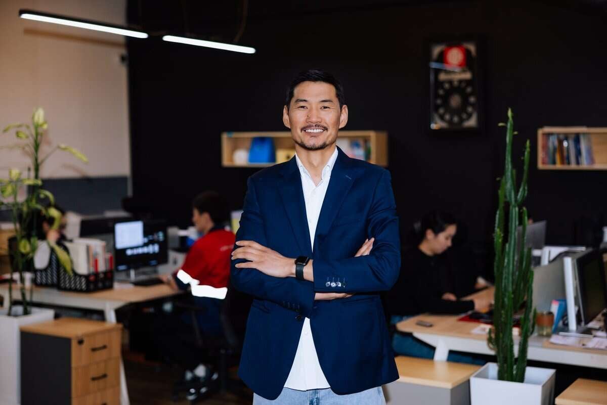 Цэнхэр пиджак өмссөн эр орчин үеийн оффис дээр гараа зөрүүлэн өөртөө итгэлтэй зогсож байна. Хүмүүс ширээн дээр компьютертэй ажилладаг, Сэтгэл шингэсэн ажилдаа. Орон зайг ургамал, тавиураар чимэглэж, урам зоригтой уур амьсгалыг бүрдүүлдэг. - Business.MN
