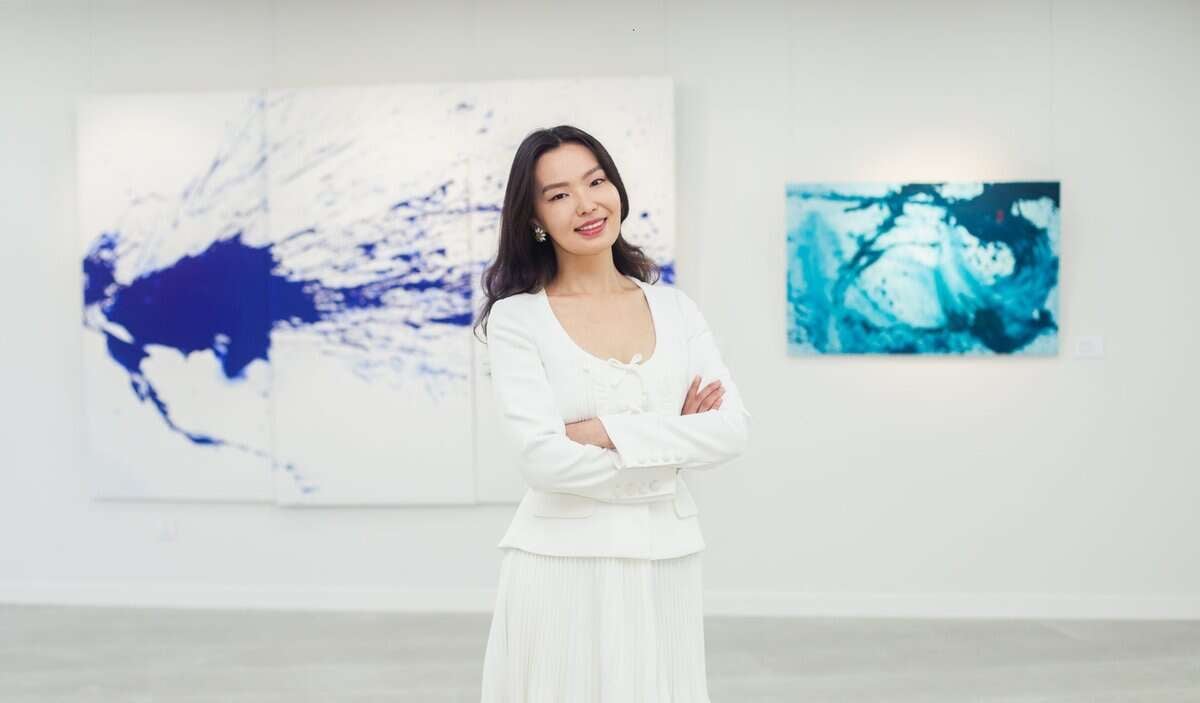 Уран зургийн галерейд цагаан хувцастай эмэгтэй гараа зөрүүлэн зогсож байгаа нь урлагийн дэгжин байдлыг илэрхийлдэг. Тэр ханыг гэрэлтүүлсэн цэнхэр өнгийн хийсвэр зургуудаар хүрээлэгдсэн инээмсэглэж байна. - Business.MN