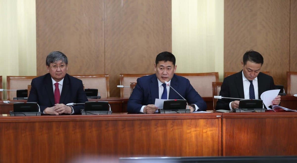 Албан ёсны хурлын танхимд гурван эрэгтэй ширээнд сууж байна. Төвд байгаа хүн, магадгүй энтрепренер байж магадгүй, цаас бариад микрофон руу ярьж байна. Хоёр талынх нь эрчүүд бичиг баримтаа харж байна. Тэд бүгд компанийнхаа эрх ашгийг төлөөлөн бизнесийн костюм өмссөн байна. - Business.MN