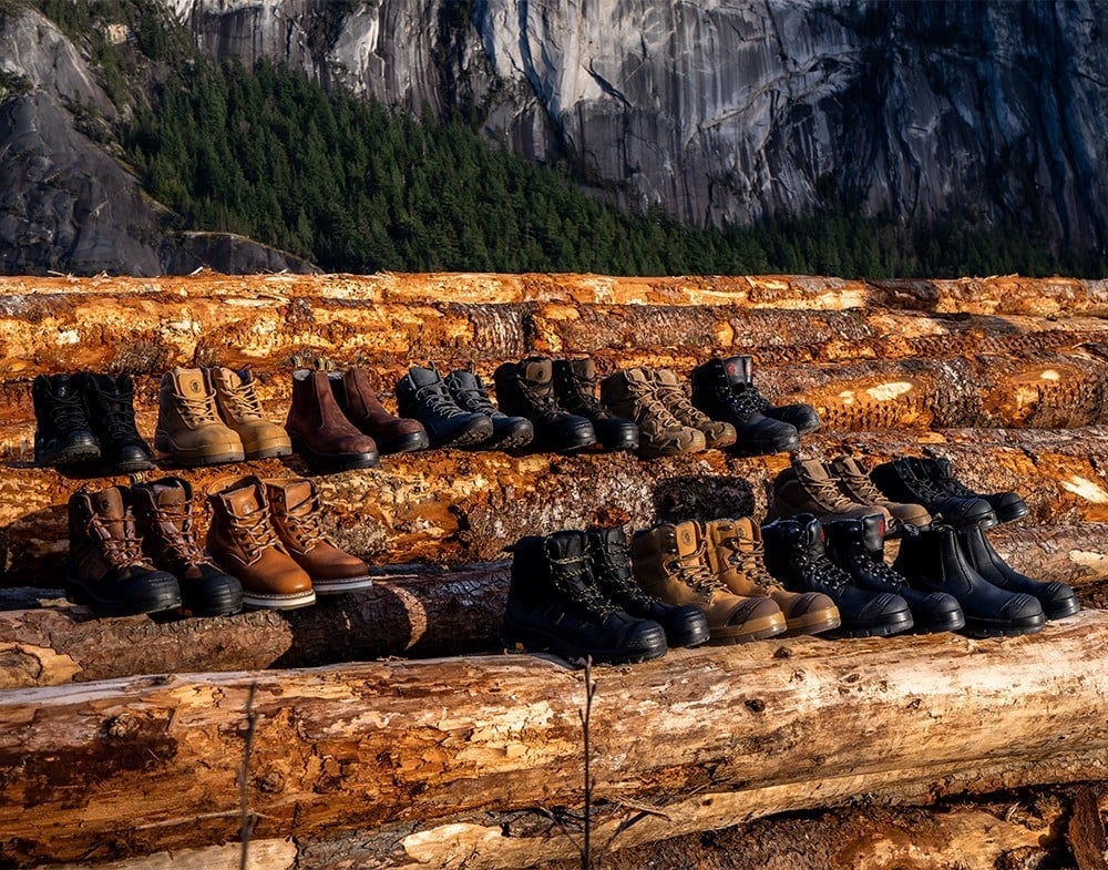 A variety of rugged work boots are lined up on large, stacked logs with a forest and rocky mountain cliff in the background, under natural daylight.