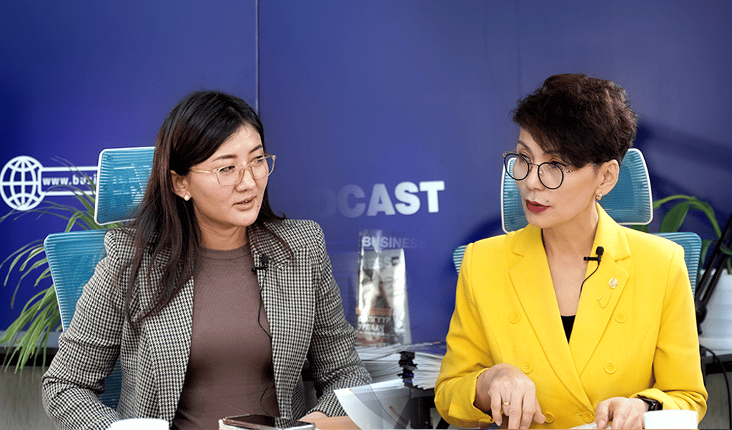 Цэнхэр, нил ягаан өнгийн дэвсгэр дээр "подкаст" гэсэн үг хэсэгчлэн харагдахуйц хоёр эмэгтэй бизнесийн талаар ярилцаж буй ширээний ард сууж байна. Нэг эмэгтэй нүдний шил, саарал алаг пиджак өмсдөг бол нөгөө нь нүдний шил, тод шар өнгийн пиджак өмсдөг. - Business.MN