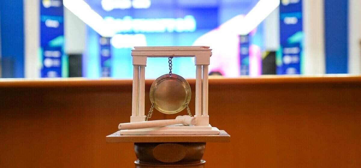 A small gong suspended in a white frame with a mallet resting beside it. It is placed on a wooden surface, with blurred digital screens displaying stock market data in the background.